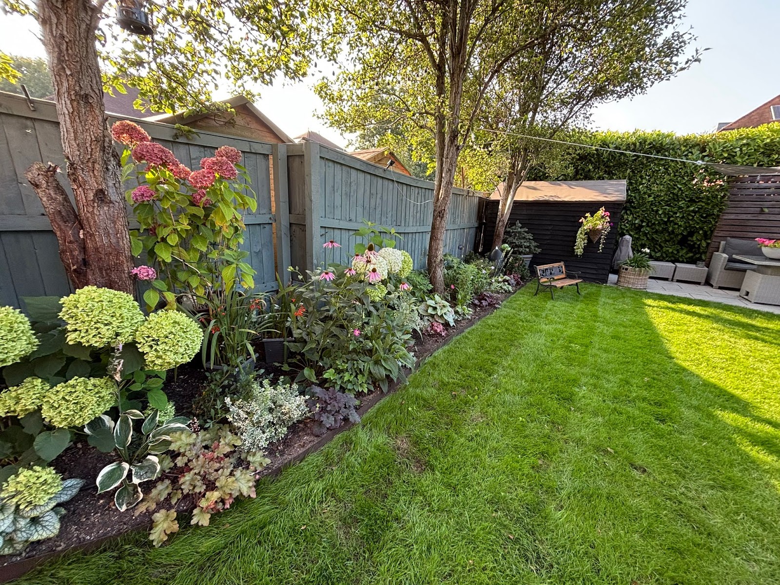 A curved brick-paved terrace with a dining set and parasol, wrapped by a low brick retaining wall and richly planted borders of salvia, heuchera and hydrangea, opening onto a mature lawn.